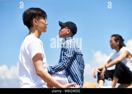 Groupe de jeunes hommes adultes asiatique assis sur des roches contre ciel bleu profitant du soleil et de l'air frais Banque D'Images