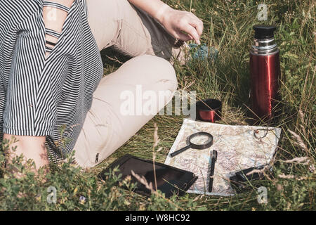 Meilleur jeune fille dans un T-shirt à rayures et pantalon brun est assis sur l'herbe. À proximité se trouve une carte, et sur c'est un comprimé, loupe, jumelles, stylo. Banque D'Images