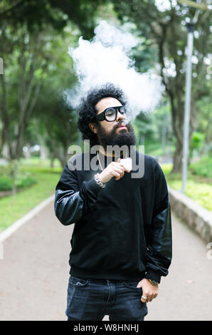 Vape. Jeune homme brutal avec grande barbe et lunettes à la mode coupe de cheveux dans un tabac cigarette électronique dans le parc de la ville. Nuage de vapeur. Style de vie. Banque D'Images