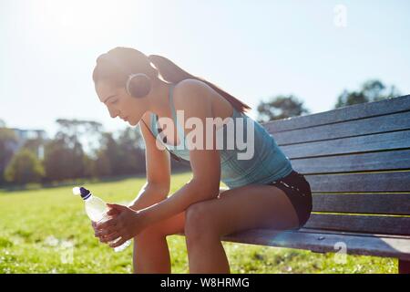 Jeune femme portant des écouteurs assis sur un banc avec une bouteille d'eau. Banque D'Images