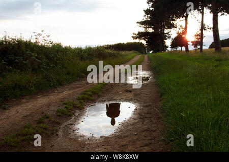 La silhouette fantomatique d'une personne qui n'est pas là, ce qui se reflète dans une flaque d'eau sur un chemin de pays au coucher du soleil. Banque D'Images