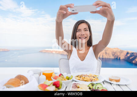 Woman taking photo de petit-déjeuner à l'aide de mobile cell smart phone app. Fille de prendre des photos d'aliments sur vacances voyages de luxe sur les médias sociaux. Belle femme en station à Santorin, Grèce, Europe. Banque D'Images