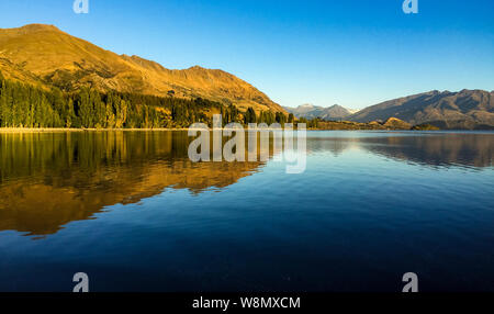 Une vue de bas niveau du lac Wanaka tôt le matin quand le soleil se lève sur les collines au loin, qui se reflètent dans les eaux scintillantes de la Banque D'Images