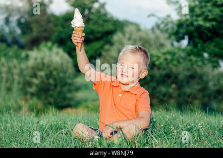 Cute smiling white Caucasian blonde garçon d'âge préscolaire en polo orange rouge holding ice cream en cornet gaufré. Kid enfant de manger des aliments en été congelé savoureux Banque D'Images