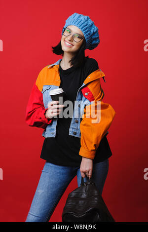 Cheerful brunette woman dans des vêtements chauds et des boissons à emporter permanent sac à dos sur fond rouge Banque D'Images