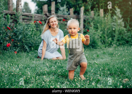 Première année jalon de l'enfant. Tout-petit bébé d'un an de faire ses premiers pas. Mère de race blanche à regarder son fils garçon marchant à l'extérieur dans le parc à l'herbe o Banque D'Images