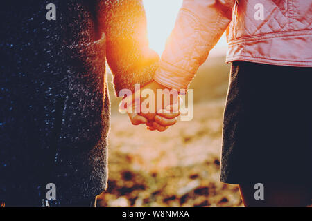 Vue arrière de la jeune homme et femme tenant la main en se tenant sur le fond flou de campagne pendant le coucher du soleil.Petit couple holding hands durin Banque D'Images