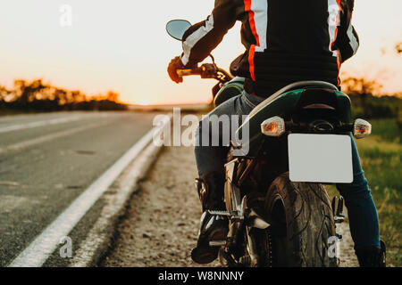 Vue arrière de la récolte de mâles en bottes et de protection rider jacket déménagement sur l'autoroute à partir de la route sur l'arrière-plan flou rétroéclairé de route vide Banque D'Images