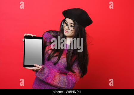 Jolie jeune femme en costume élégant et verres de sourire et démontré avec tablette moderne d'écran vide en se tenant sur le fond rouge brillant Banque D'Images