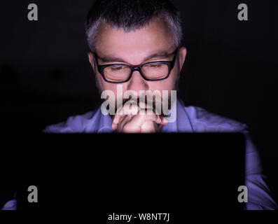 Puzzled man looking at laptop screen in dark room Banque D'Images