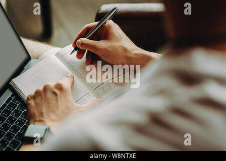 Vue arrière de la récolte de l'homme en chemise légère tenir pen et montre de l'index à la page d'éphéméride de pointage à l'ordinateur Banque D'Images