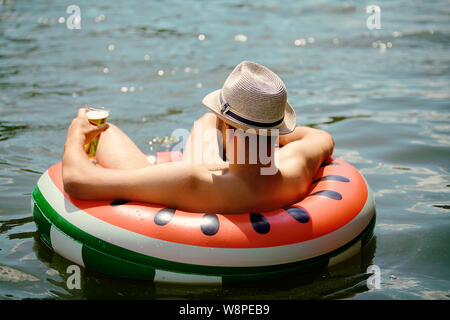 Young caucasian man with hat sur sa tête est à la dérive sur l'anneau gonflable tout en vous relaxant sur la nature, il y a un verre à la main. Banque D'Images