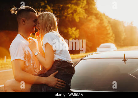 Vue latérale d'un beau jeune homme embrassant gorgeous blonde woman sitting on car les deux rient au coucher du soleil sur fond rétroéclairé de road Banque D'Images