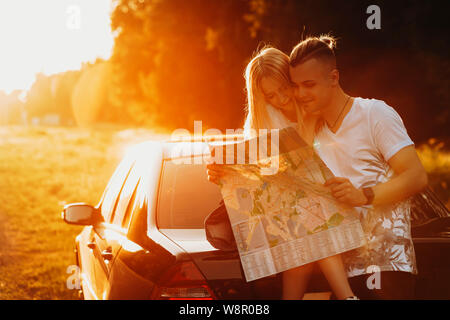 Happy young smiling couple un examen de près de grande carte papier avec woman sitting on car trunk au coucher du soleil sur fond pays rétroéclairé Banque D'Images