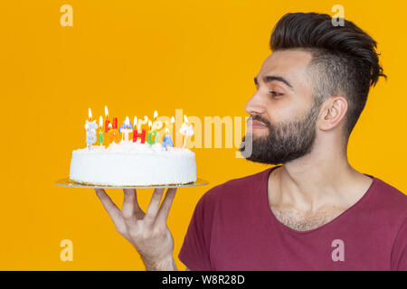 Vue latérale d'un beau jeune homme barbu arabe hors de soufflage des bougies avec un gâteau de félicitations posant sur un fond jaune Banque D'Images