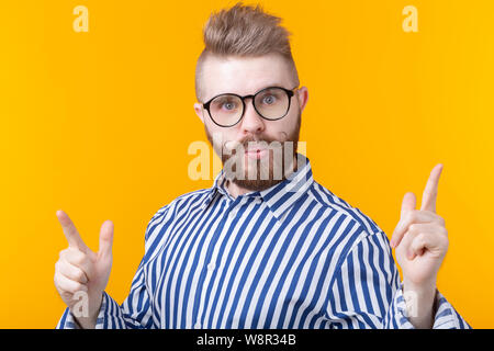 Jeune homme positif avec une moustache et barbe points heureusement son doigt vers le haut montrant quelque chose d'utile sur un fond jaune. Concept d'important Banque D'Images