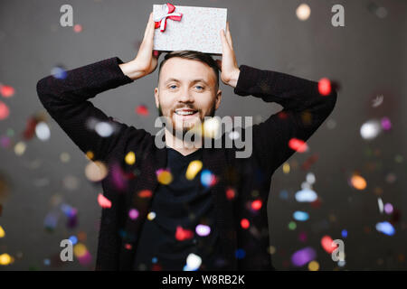 Handsome man smiling at camera et en maintenant joliment emballé présent sur la tête et de l'article derrière la chute de confettis sur fond gris Banque D'Images