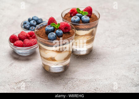 Dessert tiramisu classique avec des bleuets et framboises dans un verre et des bols de fruits rouges sur fond de béton Banque D'Images