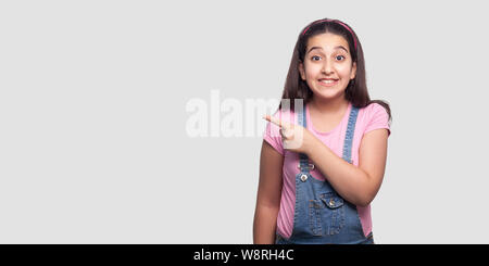 Portrait of happy beautiful brunette woman in Casual t-shirt rose et salopette bleue, permanent à pleines dents avec sourire et pointant à vide wal Banque D'Images