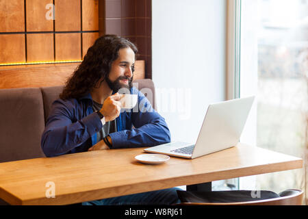 Portrait de satisfait bel homme jeunes adultes renseignement freelancer dans style casual assis dans un café avec un ordinateur portable, tasse de café potable et sourire, Banque D'Images