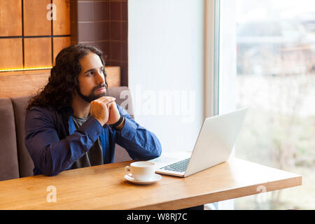 Portrait of handsome young adult man freelancer dans style casual assis dans un café avec un ordinateur portable, à la fenêtre et de la planification de son travail, bussi Banque D'Images