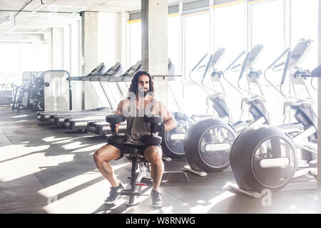 Portrait de jeune adulte construit musculaire beau sportif avec de longs cheveux bouclés travaillant dans une salle de sport, assis sur une machine d'haltérophilie et tenant deux Banque D'Images