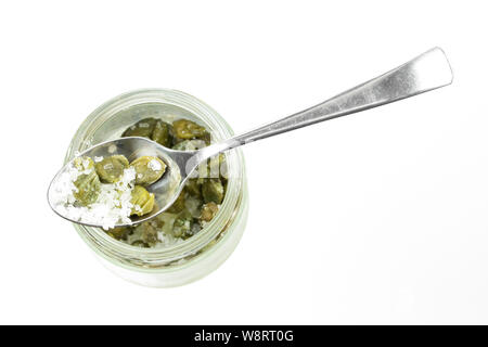 Câpres vert avec du gros sel dans une cuillère à thé sur un bocal de verre isolé sur un fond blanc, vue du dessus. Les légumes salés assaisonnement épices, ingrédients culinaires Banque D'Images