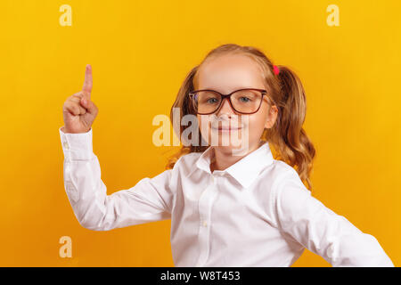 Un enfant avec des lunettes montre son index vers le haut. Portrait d'une petite fille sur un fond jaune. L'éducation et de l'école concept. Banque D'Images