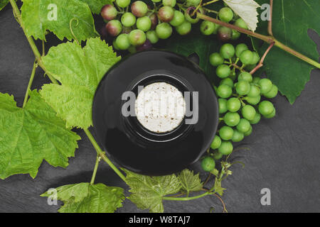 Vue supérieure de la composition de la bouteille de vin blanc et noir avec du liège entouré par de jeunes rameaux de raisin avec des feuilles vertes et les raisins ne sont pas mûrs sur pierre noire Banque D'Images