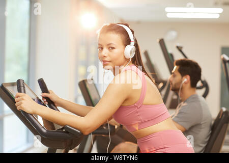 Waist up portrait of young woman wearing headphones looking at camera tout en exerçant en salle de sport, copy space Banque D'Images