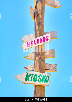 Grand poster avec panneaux en bois couleur pointant vers des destinations diverses et une plage services en espagnol sur un ciel bleu. Kiosque et des glaces. Banque D'Images