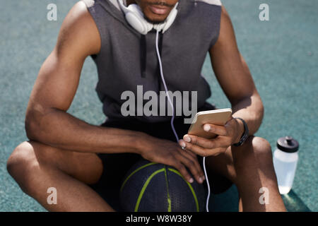 La mi-section Fond d'Afro-américain contemporain man using smartphone tout en étant assis sur un terrain de basket-ball, copy space Banque D'Images