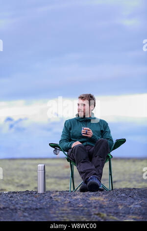 Homme assis dans Camping chaise pliante de boire du café à partir de la bouteille thermos flask au crépuscule dans la nature sur l'Islande. Camping-pensée relaxant pensive de prendre de pause dans road trip in belle la nature islandaise. Banque D'Images