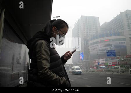 Un passager portant un masque de visage utilise son téléphone portable à une station de bus dans le smog lourde à Beijing, Chine, 27 mars 2014. Le smog épais a continué d'affec Banque D'Images