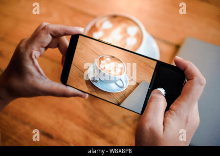 Part de prendre une photo de la tasse de café latte art avec l'art de la mousse sur table en bois avec son smart phone assis à un café et partager snap on social Banque D'Images