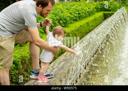 Papa se promène avec sa petite fille dans le parc avec des fontaines. Baby Girl 10 mois tire les mains pour des jets d'eau dans une fontaine European Caucasian baby Banque D'Images