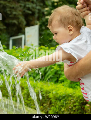 Baby Girl étend sa main vers la fontaine, le bébé joue avec jets d'eau dans le parc. Un enfant sur une promenade dans le parc joue avec de l'eau dans un puits Banque D'Images