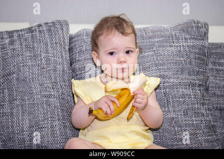 La petite fille de 10 mois mange une banane mûre. Un petit enfant mange des fruits exotiques. Fille en jaune avec une banane dans ses mains. Une petite fille Banque D'Images