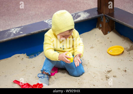 Bébé Enfant fille garçon d'un an à jouer sur l'aire de jeux dans le bac à sable. Un enfant en jeans et vêtements jaune se trouve dans un bac à sable et joue avec le sable m Banque D'Images