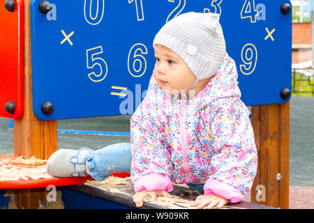 Petite fille en une veste et un chapeau monte dans le bac à sable, jette son pied sur le côté du bac à sable. Fille sur une promenade sur l'aire de jeu avec san Banque D'Images