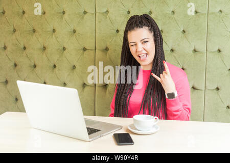 Portrait of attractive young girl indépendant avec des dreadlocks hairstyle en chemisier rose assis dans un café, travailler sur ordinateur portable et clignant montrant roc Banque D'Images