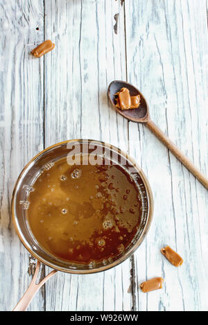 Casserole de sauce caramel fondu avec une cuillère de bois et morceaux de bonbons sur une table en bois blanc arrière-plan. Banque D'Images