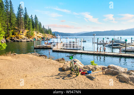 Un scooter et vélo s'asseoir sur les rives du lac de Coeur d'Alene resort près de la marina, un groupe d'adolescents marchant le long de la promenade passé amarré boats. Banque D'Images