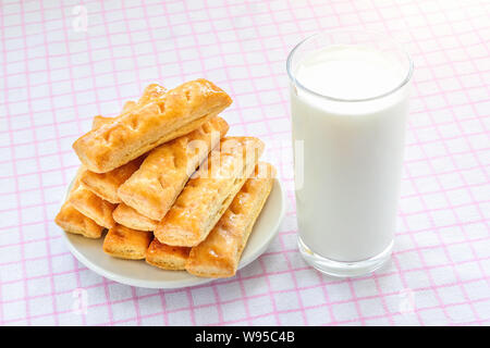 Verre de lait de vache pâte feuilletée sucré et les cookies sur une soucoupe blanche sur fond blanc à carreaux rose nappe. Délicieux petit-déjeuner avec du lait. Banque D'Images