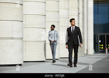 Confident businessman sérieux en costume noir à marcher le long de la rue de la ville avec l'autre homme dans l'arrière-plan Banque D'Images