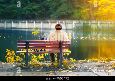 Femme solitaire âgée avec un petit chien noir assis sur un banc près du lac ou de la rivière d'automne dans un parc de la ville. Détente. Concept de solitude Banque D'Images