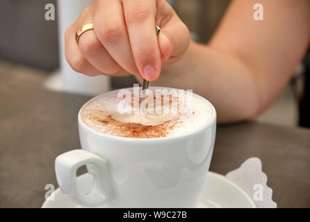 Libre de la main d'une femme avec une bague de mariage en remuant son cappuccino dans une tasse blanche Banque D'Images