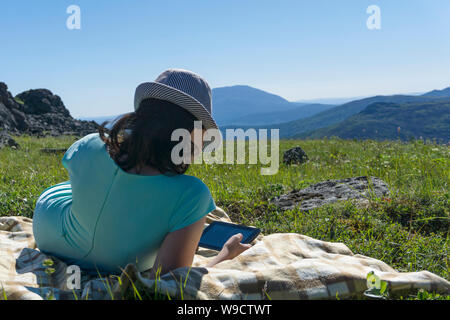 Fille en robe et chapeau assis sur une couverture avec une tablette dans ses mains sur une prairie alpine sur un fond de montagnes, la vue de l'arrière Banque D'Images