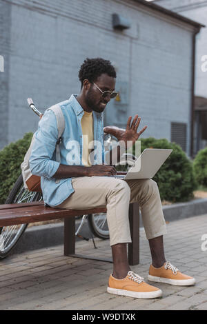 Cheerful african american man en agitant la main et assis sur un banc pendant l'appel vidéo dans coffre Banque D'Images