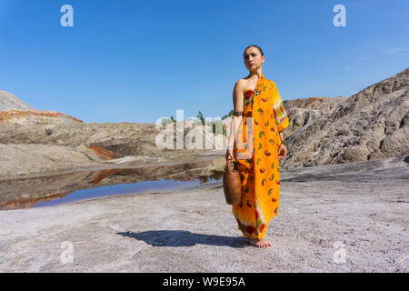 Jeune femme en robe ethnique pour marcher avec de l'eau pot en céramique dans paysage de désert Banque D'Images